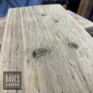 Puede incluir: Una tabla de madera rectangular de color claro y sin acabado, con un patr&oacute;n de veta prominente, nudos visibles y textura superficial natural. Un logotipo cuadrado en la esquina inferior izquierda muestra el texto DAVES HARDWOOD en blanco.