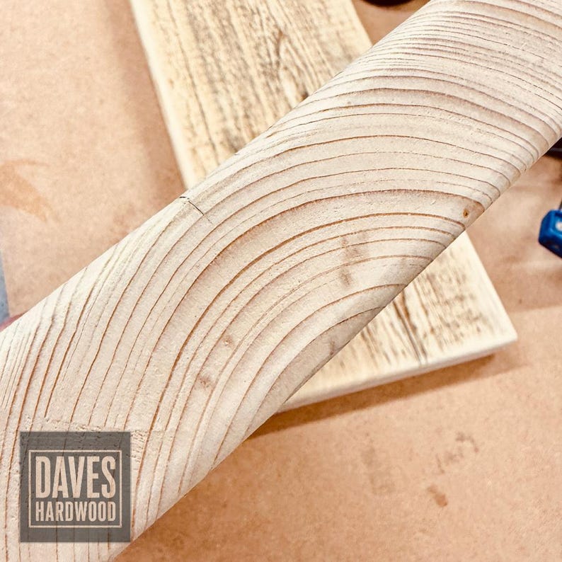 Puede incluir: Una tabla de madera de color claro con patrones de veta curvos y pronunciados y una textura suave. Un logotipo cuadrado en la esquina inferior izquierda muestra el texto DAVES HARDWOOD en una fuente sans-serif negrita, indicando el origen.