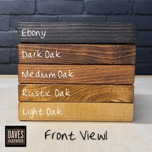 Puede incluir: Una pila de cinco tablas de madera rectangulares con diferentes acabados de tinte. Las etiquetas Ebony, Dark Oak, Medium Oak, Rustic Oak y Light Oak est&aacute;n escritas en cada tabla. El logotipo de Daves Hardwood y el texto Front View son visibles.