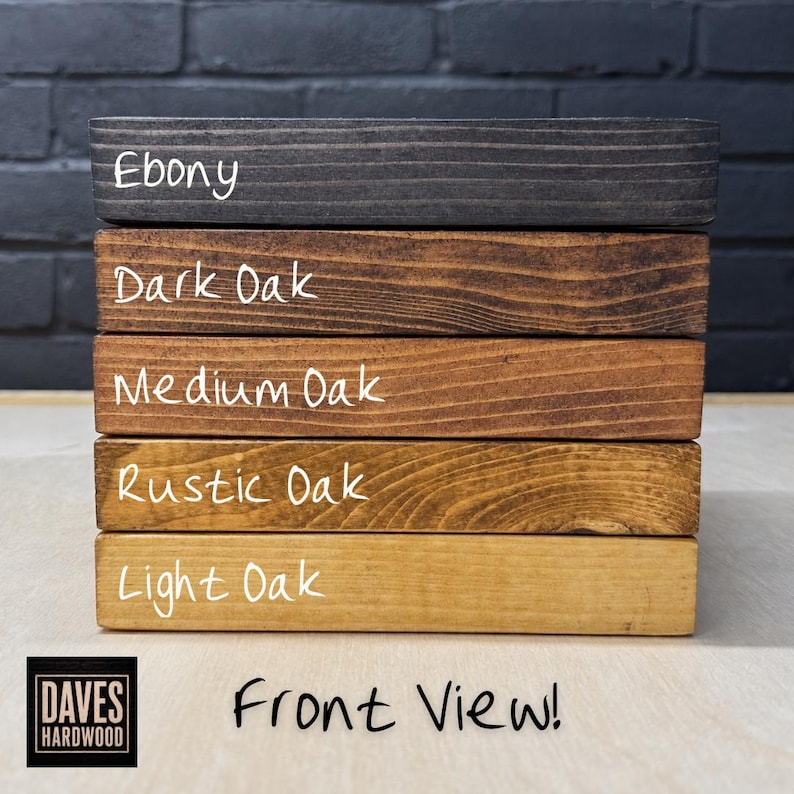 Puede incluir: Una pila de cinco tablas de madera rectangulares con diferentes acabados de tinte. Las etiquetas Ebony, Dark Oak, Medium Oak, Rustic Oak y Light Oak est&aacute;n escritas en cada tabla. El logotipo de Daves Hardwood y el texto Front View son visibles.