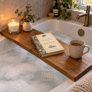 Puede incluir: Una bandeja rectangular de madera marrón oscuro para bañera colocada sobre una bañera blanca llena de agua jabonosa. La bandeja sostiene una vela encendida en un frasco de vidrio, una pequeña planta en maceta, un libro titulado The Little History of Essex.
