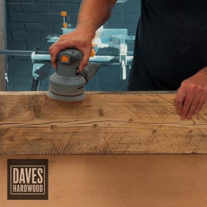 Puede incluir: Una lijadora orbital el&eacute;ctrica gris se utiliza para alisar la superficie de una pieza larga y rectangular de madera marr&oacute;n natural con vetas y nudos visibles. El logotipo de Daves Hardwood est&aacute; impreso en la esquina inferior izquierda.