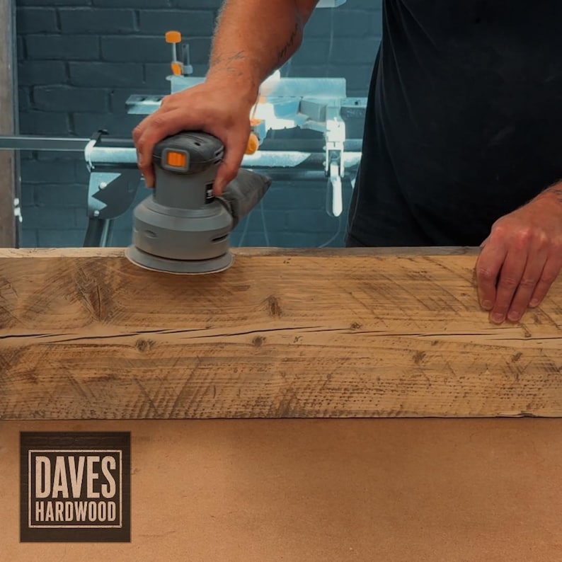 Puede incluir: Una lijadora orbital el&eacute;ctrica gris se utiliza para alisar la superficie de una pieza larga y rectangular de madera marr&oacute;n natural con vetas y nudos visibles. El logotipo de Daves Hardwood est&aacute; impreso en la esquina inferior izquierda.