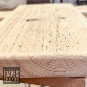 Puede incluir: Una pieza rectangular de madera de color claro con un patr&oacute;n de veta visible y un nudo oscuro. La superficie muestra una textura natural y tonos marr&oacute;n claro. Un logotipo cuadrado en la esquina inferior izquierda contiene el texto DAVES HARDWOOD.