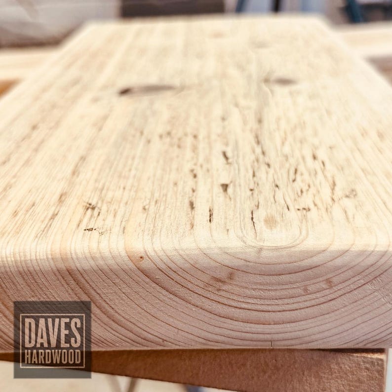 Puede incluir: Una pieza rectangular de madera de color claro con un patr&oacute;n de veta visible y un nudo oscuro. La superficie muestra una textura natural y tonos marr&oacute;n claro. Un logotipo cuadrado en la esquina inferior izquierda contiene el texto DAVES HARDWOOD.