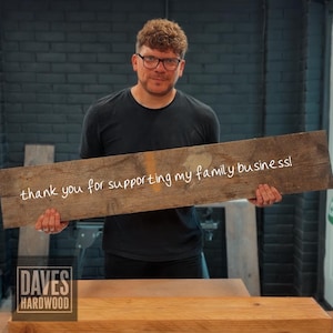 Puede incluir: Una pieza rectangular de madera marr&oacute;n desgastada sostenida horizontalmente, con el texto escrito a mano en blanco "thank you for supporting my family business!" en el centro, con veta de madera visible y textura r&uacute;stica.