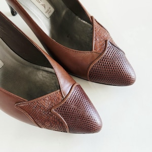 May include: A pair of brown leather pumps featuring a pointed toe with a dark brown textured snakeskin-patterned overlay. The interior lining is a light grey color, and the heel is visible at the back. The brand label inside reads LISA MADE IN ITALY.