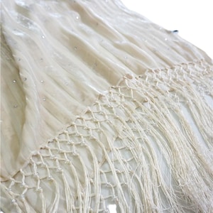 May include: A cream-colored velvet shawl featuring a subtle crinkled texture and scattered small clear rhinestones. The bottom edge is finished with a decorative knotted macrame fringe pattern that transitions into long, flowing loose threads.