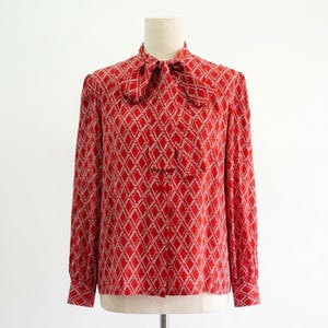 May include: A long-sleeved red blouse featuring a white diamond lattice pattern throughout the fabric. The garment includes a high neckline with an integrated tie-front bow detail, long sleeves with buttoned cuffs, and a straight hemline.