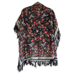May include: A black shawl featuring a vibrant floral pattern with red, purple, and white flowers and green leaves. The garment has a grey border along the edges and is finished with long, black fringe detailing along the bottom hemline.