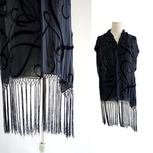 May include: A black sleeveless wrap or shawl featuring a sheer fabric base with a raised black velvet swirl pattern throughout. The garment has a straight hemline finished with long, thin black fringe detailing that hangs from a knotted mesh border.