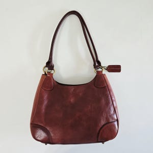 May include: A dark reddish-brown leather shoulder bag featuring two thin straps attached to gold-toned metal rings. The bag has a structured base with reinforced corners and a small rectangular leather tag hanging from one of the side rings.