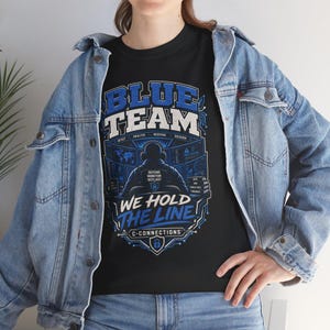 May include: A black t-shirt featuring a graphic of a person wearing headphones in front of computer monitors. The text reads BLUE TEAM, WE HOLD THE LINE, C-CONNECTIONS, DEFEND MONITOR OUTLAST, DETECT, ANALYZE, RESPOND, RECOVER. The shirt is worn under a light blue denim jacket.