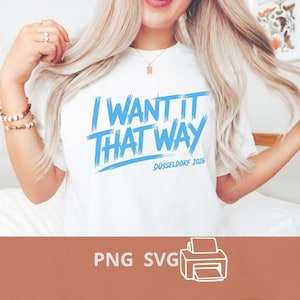 Puede incluir: Una camiseta blanca de manga corta con el texto azul I WANT IT THAT WAY y DÜSSELDORF 2026 en un estilo de fuente de pincelada. El diseño está centrado en el pecho de la prenda, que tiene un cuello redondo y un ajuste estándar.