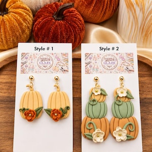 May include: Two pairs of dangle earrings on white display cards labeled Glitz & Glaze. Style 1 features single light orange pumpkins with orange flowers and green leaves. Style 2 features stacked pumpkins in orange, green, and tan with white flowers.