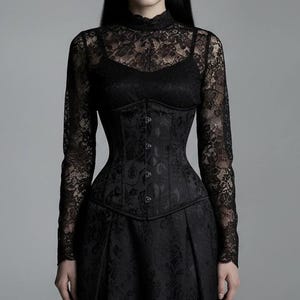 Peut inclure: Une robe noire à manches longues dotée d'un haut en dentelle à col montant avec des motifs floraux complexes. Le corsage comprend une section centrale structurée style corset avec des coutures verticales et une rangée de boutons.