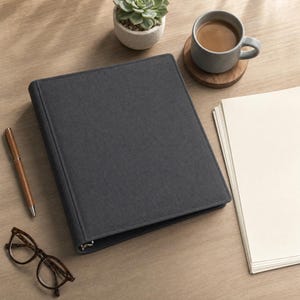 Op de afbeelding: Een donkergrijze, met stof beklede ordner met drie ringen en een textuurafwerking. De ordner ligt naast een bruine pen, een bril met schildpadpatroon, een grijze keramische mok gevuld met koffie en een stapel blanco wit papier.