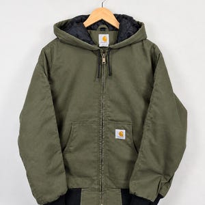May include: An olive green hooded jacket with a full-length front zip, black quilted interior lining, and black ribbed cuffs at the waist and sleeves. The jacket features a Carhartt brand label on the chest pocket and the inner neck collar.