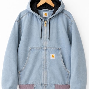 Puede incluir: Una chaqueta con capucha y cremallera en tejido vaquero azul claro, con forro acolchado negro, cremallera frontal en tono latón y puños y dobladillo acanalados en un tono malva apagado. El logotipo de Carhartt aparece en el pecho y el cuello.