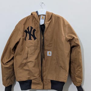 May include: A tan hooded jacket featuring a black New York Yankees logo embroidered on the right chest. The jacket has a full-length front zipper, black ribbed cuffs and hem, and a Carhartt brand patch on the front left pocket area.