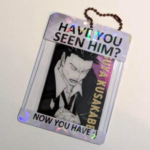 Peut inclure: Un porte-carte rectangulaire en plastique holographique présentant une illustration en noir et blanc de style anime. Le texte HAVE YOU SEEN HIM? est imprimé en haut et NOW YOU HAVE :) en bas. Une chaînette à billes marron est fixée.