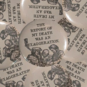 May include: A circular pin-back button featuring a black and white illustration of two figures and a skeleton. The text printed on the button reads, THE REPORT OF MY DEATH WAS AN EXAGGERATION. The design is repeated across multiple overlapping buttons.