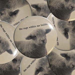 May include: A circular pinback button featuring a black and white halftone image of a lamb. The text "the rage within me builds." is printed in a black typewriter-style font above the lamb's head. The design is repeated across multiple buttons.