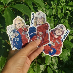 May include: Three die-cut stickers featuring illustrated hockey players in red hoodies. The left sticker shows Desbiens with number 35, the centre shows Poulin with number 29, and the right shows Roque with number 26. Each sticker includes a name and year.