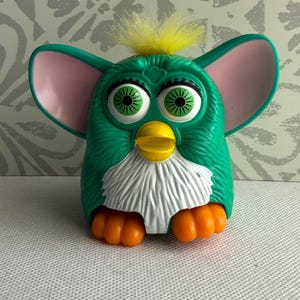 May include: A green electronic toy with large pink-lined ears, bright green eyes, a yellow beak, and orange feet. The toy features a white chest patch and a tuft of yellow hair on top of its head, with a molded plastic body and textured fur details.
