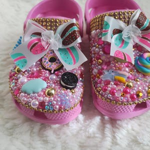 May include: A pair of pink clogs decorated with gold rhinestones, assorted pink and white pearls, and various charms including a donut, a cookie, a blue cloud, a shooting star, and a colorful swirl. Each shoe features a large white bow.