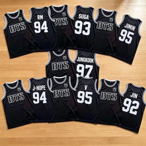Op de afbeelding: Een collectie zwarte basketbalshirts met witte biezen en witte zeefdruktekst. Sommige shirts tonen de letters BTS, terwijl andere namen zoals RM, SUGA, JIMIN, JUNGKOOK, J-HOPE, V en JIN bevatten, gecombineerd met de geboortejaren.