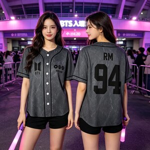 Pode incluir: Uma camisa de beisebol cinza-escura com padrão marmorizado, acabamentos pretos, botões brancos e o texto BTS e RM 94 impresso na frente e nas costas. A peça de manga curta é combinada com calções pretos, exibindo um design de inspiração esportiva.