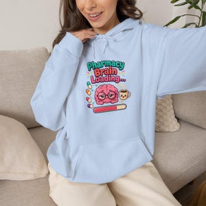 May include: A light blue hooded sweatshirt featuring a graphic print of a pink brain wearing glasses, a coffee mug, and colorful pills. The text reads Pharmacy Brain Loading... above a loading bar. The hoodie has long sleeves and a front pocket.