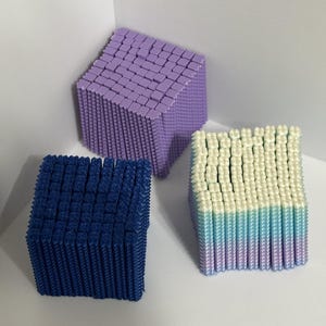 May include: Three cubic structures composed of numerous small, vertical, square-topped pillars. One cube is solid lavender, one is solid dark blue, and the third features a gradient transition from white at the top to light blue and lavender at the base.