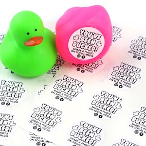 May include: A close-up of a sheet of stickers with the text "You've Been & Ducked! Share a photo on social media" printed on each sticker. There are two rubber duck toys, one green and one pink, in the background.