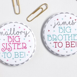 May include: Two white round buttons with gray borders and colorful confetti. One button says "mallory BIG SISTER TO BE!" in pink and blue lettering. The other button says "james BIG BROTHER TO BE!" in blue and red lettering.