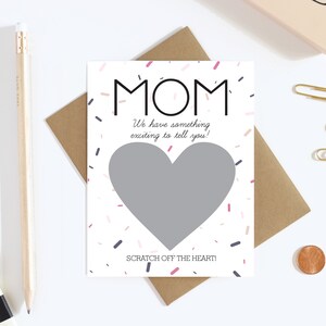 May include: A white card with the word "MOM" in black, with a gray heart-shaped scratch-off area. The card reads "We have something exciting to tell you!" and "SCRATCH OFF THE HEART!" with confetti accents.