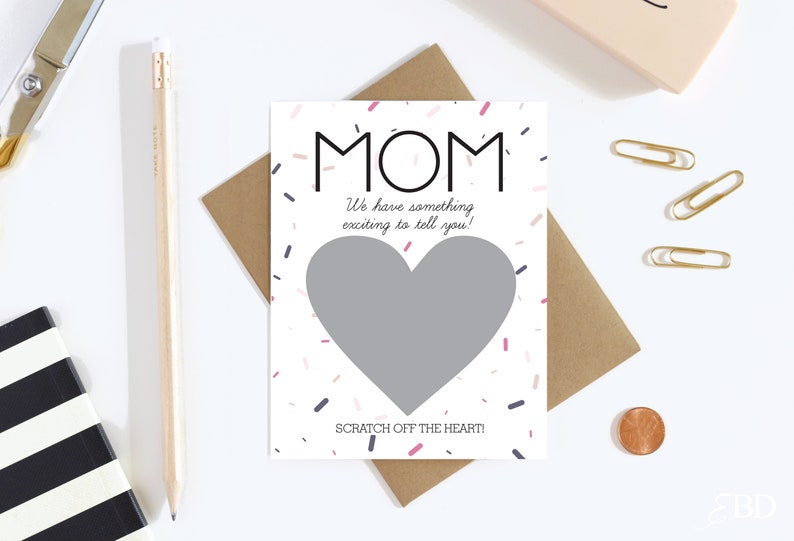 May include: A white card with the word "MOM" in black, with a gray heart-shaped scratch-off area. The card reads "We have something exciting to tell you!" and "SCRATCH OFF THE HEART!" with confetti accents.