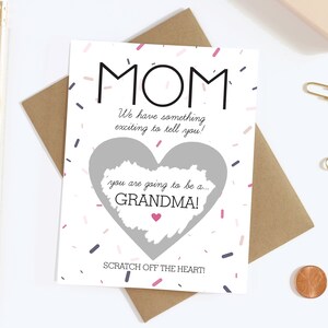 May include: A white card with the word "MOM" in black, and the text "We have something exciting to tell you!" The card has a heart-shaped scratch-off area that reveals "you are going to be a... GRANDMA!" with confetti and a pink heart.