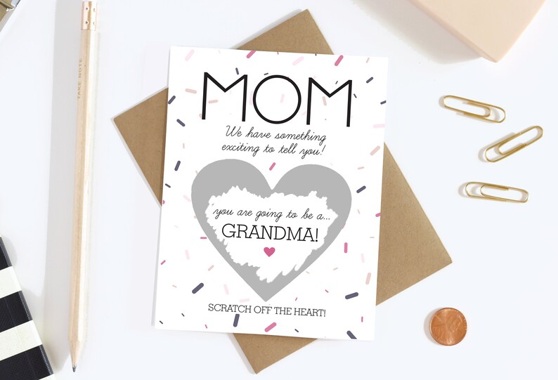 May include: A white card with the word "MOM" in black, and the text "We have something exciting to tell you!" The card has a heart-shaped scratch-off area that reveals "you are going to be a... GRANDMA!" with confetti and a pink heart.