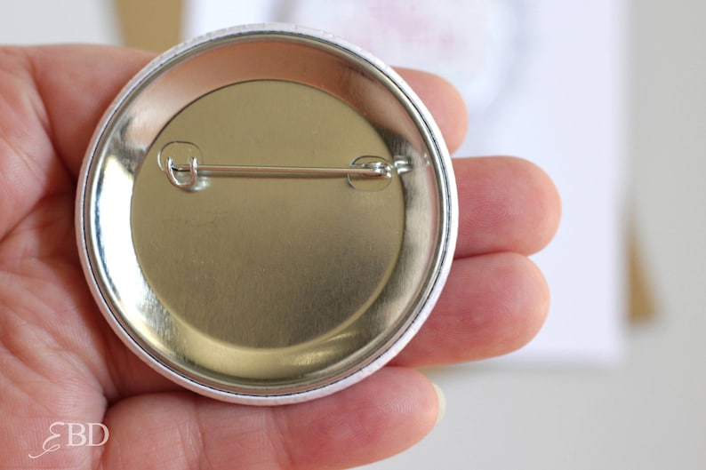 May include: The back of a white button with a silver pin and a silver metal backing.