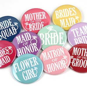 May include: Ten round buttons with different bridal party titles printed in white on colorful backgrounds. The titles include "Bride Squad", "Mother of the Bride", "Bridesmaid", "Maid of Honor", "Bride", "Team Bride", "Jr. Bridesmaid", "Flower Girl", "Matron of Honor", and "Mother of the Groom".