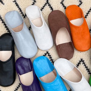 Puede incluir: Una colección de ocho zapatillas de cuero en varios colores sólidos, incluyendo negro, azul claro, blanco, marrón, naranja, morado y azul brillante. Cada zapatilla presenta una punta redondeada, un diseño sin cordones y un interior liso.