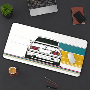 May include: A rectangular desk mat featuring a white BMW M3 car illustration viewed from the rear. The design includes horizontal teal and yellow stripes with thin red and blue lines. The car graphic shows the trunk, taillights, and M3 badge.