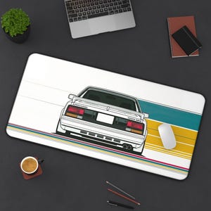 Pode incluir: Um tapete de secretária retangular com uma ilustração de um carro Toyota MR2 branco visto de trás. O design inclui riscas horizontais em azul-petróleo, amarelo e vermelho na parte inferior. O texto "SUPERCHARGED" e "TOYOTA MR2" está impresso.