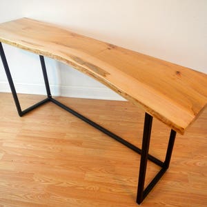 May include: A long, narrow wooden table with a live edge top and black metal legs. The table is made of light wood with a natural finish. The legs are simple and modern, and the table is perfect for a small space.