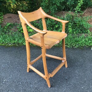 May include: A wooden bar stool with a unique, organic design. The stool has a curved back and armrests, and the legs are shaped like branches. The stool is made of light-colored wood and has a natural finish.