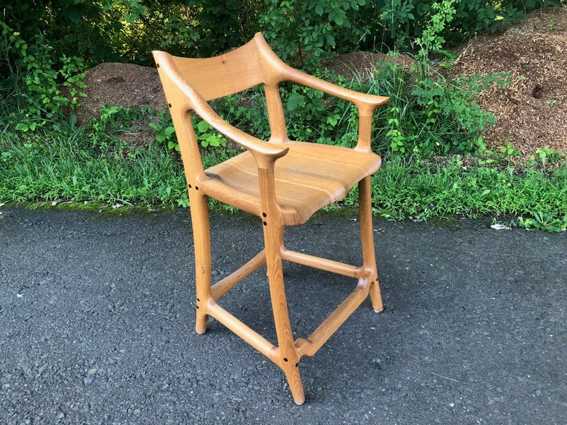 May include: A wooden bar stool with a unique, organic design. The stool has a curved back and armrests, and the legs are shaped like branches. The stool is made of light-colored wood and has a natural finish.