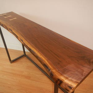 May include: A long, narrow console table with a live edge walnut top and a black metal frame. The table has a natural wood finish and a rustic look.