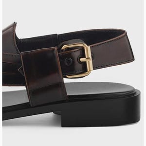 K&ouml;nnte beinhalten: Eine dunkelbraune Ledersandale mit einer gl&auml;nzenden, goldfarbenen Metallschnalle am Seitenriemen. Das Schuhwerk verf&uuml;gt &uuml;ber eine glatte, flache schwarze Sohle und ein strukturiertes Fersenriemendesign mit einer gl&auml;nzenden Lederoberfl&auml;che.